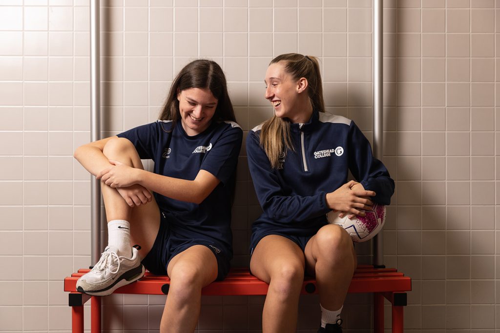 Two womens football players laughing whilst sat on a bench