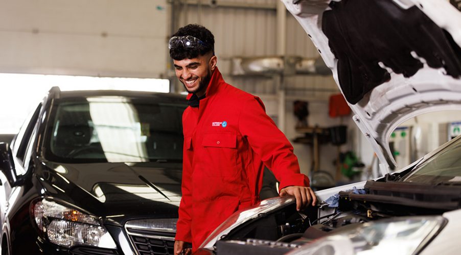 Automotive student in red overalls smiling whilst looking at car engine