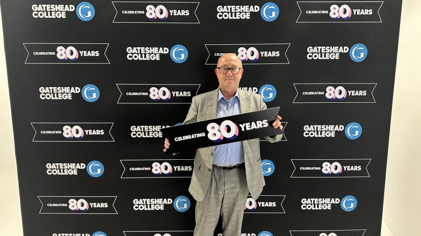 A man wearing a suit stands in front of a banner saying 'Gateshead College celebrating 80 Years'