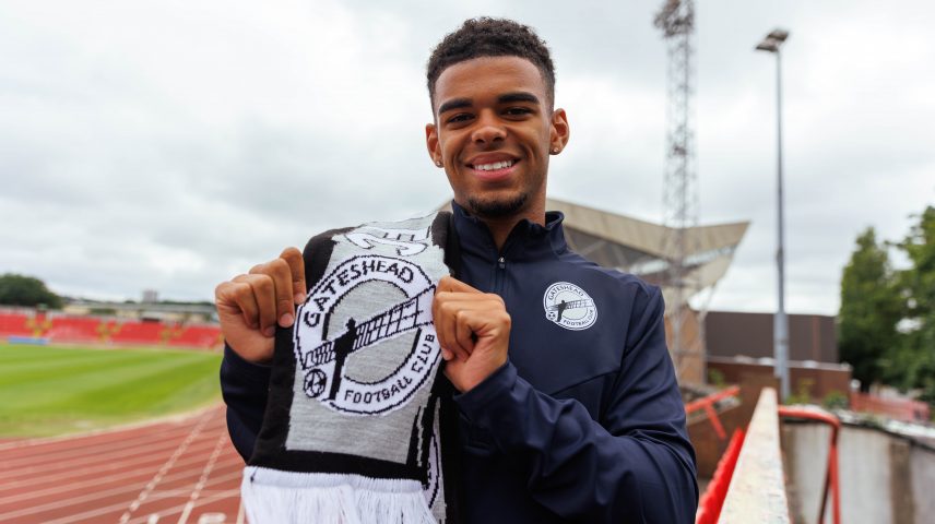 A young footballer stands on a football pitch holding a Gateshead Football Club scarf