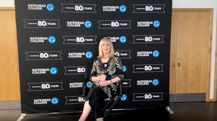 A lady sitting in front of a backdrop that says Gateshead College celebrating 80 years