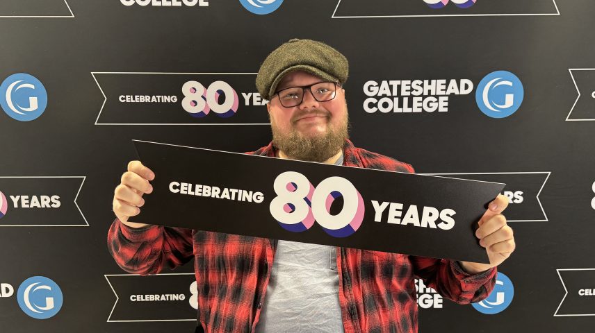 A man wearing hat and glasses sitting holding a sign saying 'celebrating 80 years'