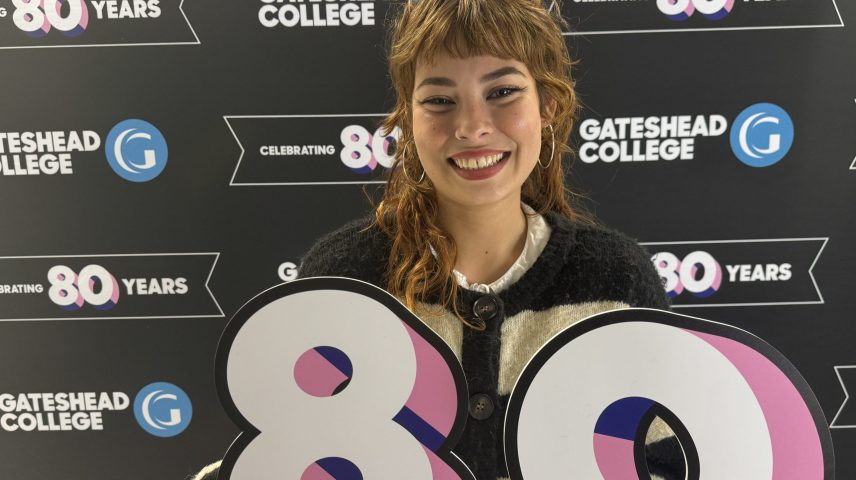 A woman smiling holding and 8 and 0 in front of a backdrop saying Gateshead College celebrating 80 years