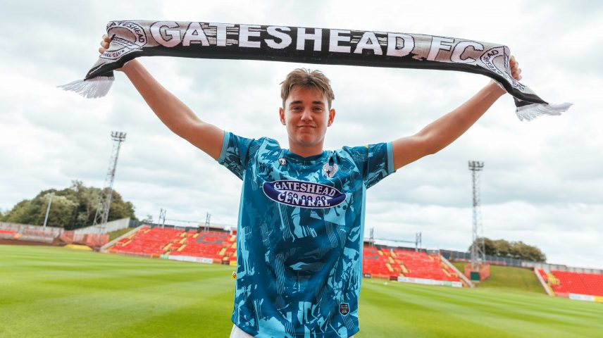 A young man stands on a football pitch holding a Gateshead FC football scarf above his head.