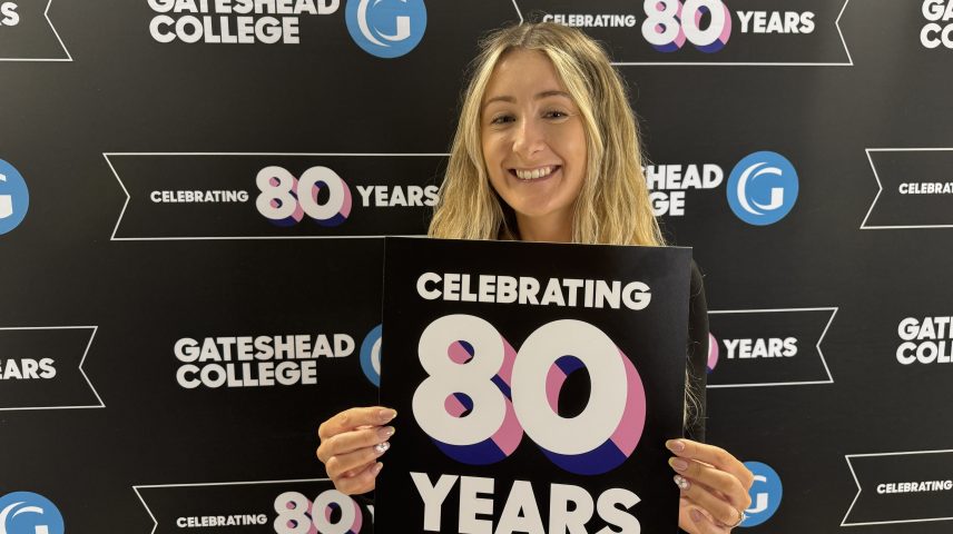 A lady with blonde hair standing infront of a Gateshead College backdrop holding a sign saying celebrating 80 years.