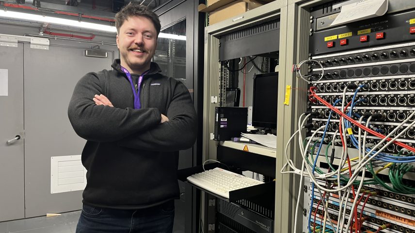 A man standing with his arms crossed standing infront of a cabinet filled with wires.