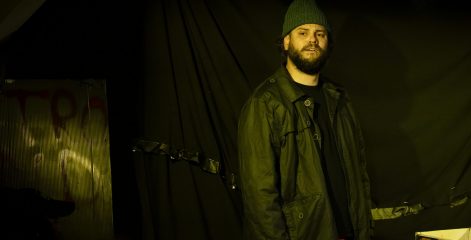 A man in a woolly hat and coat standing on a stage that's dimly lit.
